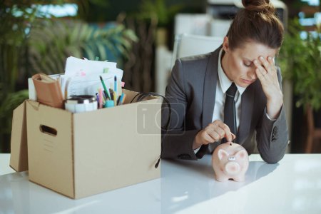 Photo for New job. stressed modern female worker in modern green office in grey business suit with personal belongings in cardboard box putting coin into piggy bank. - Royalty Free Image