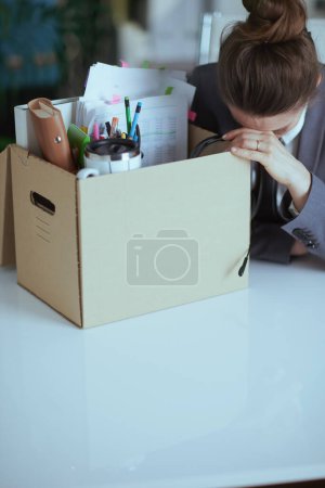 Téléchargez les photos : Un nouveau boulot. souligné femme d'âge moyen moderne employée dans un bureau vert moderne en costume d'affaires gris avec des effets personnels dans une boîte en carton. - en image libre de droit