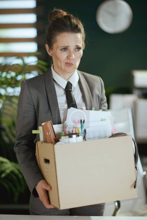Téléchargez les photos : Un nouveau boulot. travailleuse moderne confuse dans un bureau vert moderne en costume d'affaires gris avec des effets personnels dans une boîte en carton. - en image libre de droit