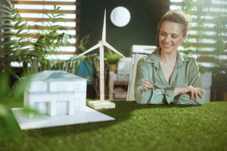 Foto de Tendencias ecológicas. feliz joven dueño de la pequeña empresa mujer en blusa verde en moderna oficina verde con casa de papel y molino de viento - Imagen libre de derechos