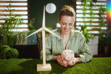 Téléchargez les photos : Tendances écologiques. femme d'affaires élégante souriante en chemisier vert dans le bureau vert moderne avec tirelire et moulin à vent - en image libre de droit