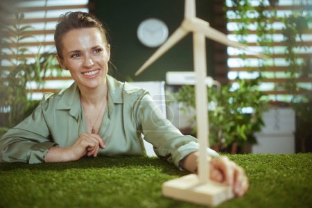 Téléchargez les photos : Des affaires durables. femme d'affaires moderne souriante en chemisier vert dans un bureau vert moderne avec moulin à vent - en image libre de droit