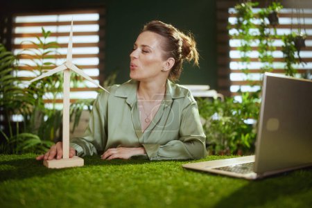 Foto de Negocio sostenible. sonriente joven mujer de negocios en blusa verde en la moderna oficina verde con ordenador portátil y molino de viento - Imagen libre de derechos