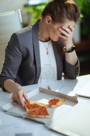 Téléchargez les photos : Un lieu de travail durable. soulignée femme comptable d'âge moyen moderne dans un costume d'affaires gris dans le bureau vert moderne avec pizza. - en image libre de droit
