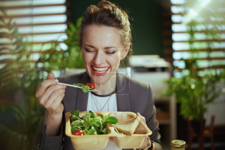 Téléchargez les photos : Un lieu de travail durable. heureuse propriétaire de petite entreprise moderne femme dans un costume d'affaires gris dans le bureau vert moderne manger de la salade. - en image libre de droit