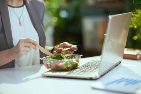 Téléchargez les photos : Un lieu de travail durable. Gros plan sur la travailleuse dans le bureau vert avec ordinateur portable manger de la salade. - en image libre de droit