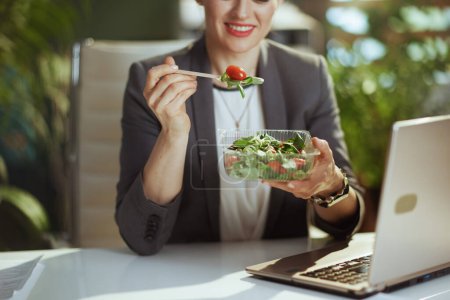 Téléchargez les photos : Un lieu de travail durable. souriant moderne 40 ans femme employée dans un costume d'affaires gris dans un bureau vert moderne avec ordinateur portable manger salade. - en image libre de droit
