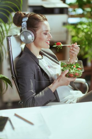 Photo for Sustainable workplace. happy modern middle aged woman worker in a grey business suit in modern green office eating salad and listening to the music with headphones. - Royalty Free Image