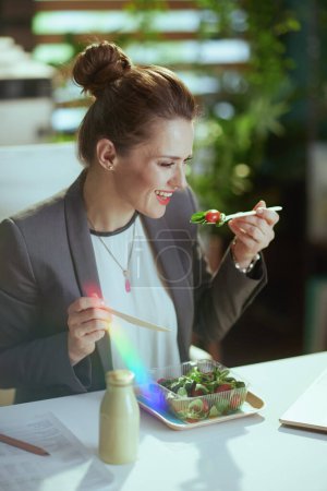 Téléchargez les photos : Un lieu de travail durable. souriant moderne 40 ans femme comptable dans un costume d'affaires gris dans le bureau vert moderne manger de la salade. - en image libre de droit