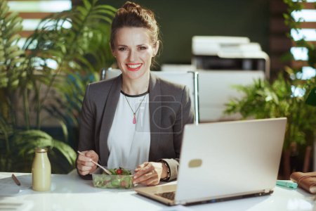 Téléchargez les photos : Un lieu de travail durable. femme comptable moderne souriante dans un costume d'affaires gris dans un bureau vert moderne avec ordinateur portable manger salade. - en image libre de droit