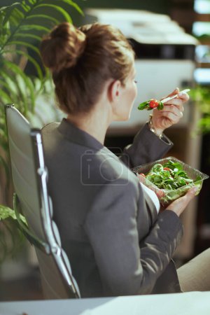 Foto de Lugar de trabajo sostenible. Visto desde atrás contable mujer en un traje gris de negocios en verde oficina comer ensalada. - Imagen libre de derechos