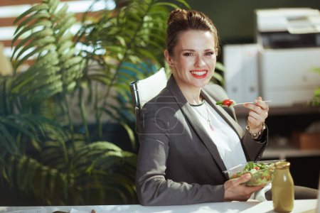 Téléchargez les photos : Un lieu de travail durable. heureuse comptable moderne femme dans un costume d'affaires gris dans le bureau vert moderne manger de la salade. - en image libre de droit