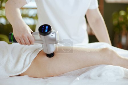 Foto de Tiempo de atención médica. Primer plano del terapeuta de masaje médico en el gabinete de masaje con pistola de masaje que masajea la pierna de los clientes. - Imagen libre de derechos
