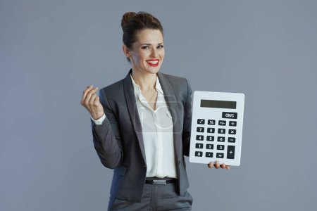 Téléchargez les photos : Heureux élégant 40 ans propriétaire de petite entreprise femme en costume gris avec calculatrice isolé sur gris. - en image libre de droit