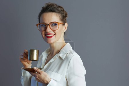 Téléchargez les photos : Heureuse travailleuse à la mode en chemisier blanc avec des lunettes et tasse de café isolé sur fond gris. - en image libre de droit