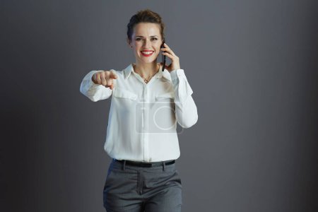 Téléchargez les photos : Heureuse propriétaire de petite entreprise d'âge moyen femme en chemisier blanc pointant à la caméra et en utilisant un smartphone isolé sur gris. - en image libre de droit