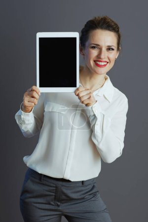 Foto de Sonriente mujer moderna de mediana edad propietaria de una pequeña empresa en blusa blanca que muestra la pantalla en blanco de la tableta PC aislada sobre fondo gris. - Imagen libre de derechos