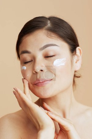 Téléchargez les photos : Femme moderne détendue avec crème faciale sur le visage isolé sur fond beige. - en image libre de droit