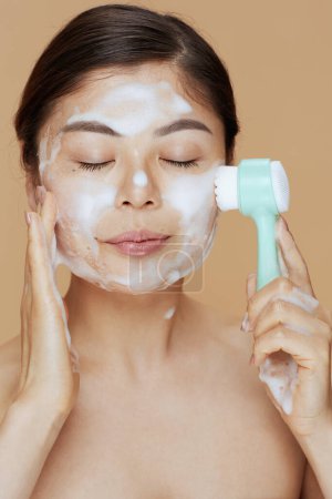 Photo for Relaxed young asian female with massager washing face on beige background. - Royalty Free Image