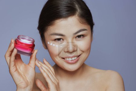Photo for Portrait of young asian woman with facial cream jar and eye cream on face against blue background. - Royalty Free Image