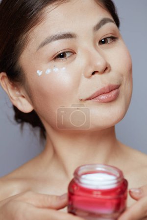 Foto de Moderna mujer asiática con crema facial tarro y crema para los ojos en la cara aislado sobre fondo azul. - Imagen libre de derechos