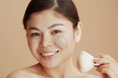 Téléchargez les photos : Portrait de jeune femme asiatique avec pinceau de maquillage sur fond beige. - en image libre de droit