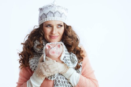 Photo for Hello winter. pensive trendy female in sweater, mittens, hat and scarf isolated on white background with piggy bank. - Royalty Free Image