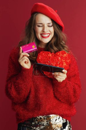 Téléchargez les photos : Joyeux Valentin. heureuse femme élégante en pull rouge et béret avec coeur rouge, smartphone et carte de crédit faire des achats en ligne sur le site e-commerce. - en image libre de droit