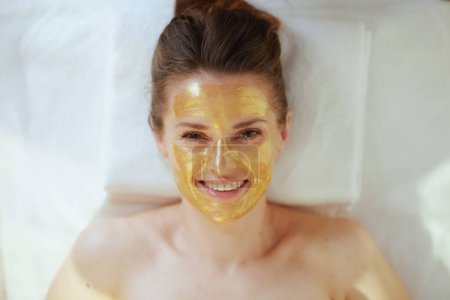 Photo for Healthcare time. Upper view of smiling modern woman in spa salon with golden cosmetic mask on face laying on massage table. - Royalty Free Image