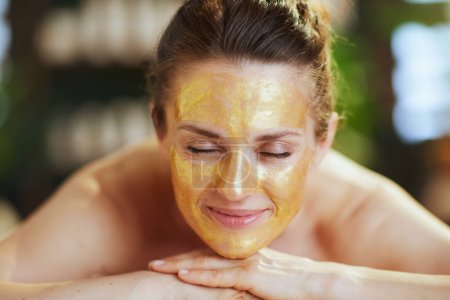Foto de Tiempo de atención médica. mujer moderna relajada en el salón de spa con máscara cosmética dorada en la cara que pone en la mesa de masaje. - Imagen libre de derechos