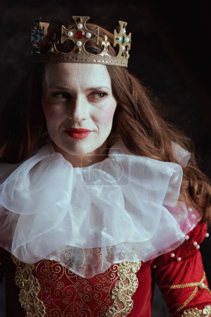 Photo for Insidious medieval queen in red dress with white collar and crown on dark gray background. - Royalty Free Image
