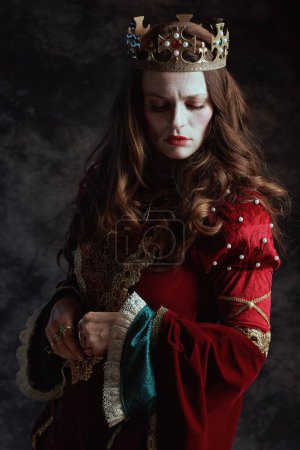 Téléchargez les photos : Triste reine médiévale en robe rouge avec maquillage blanc et couronne sur fond gris foncé. - en image libre de droit