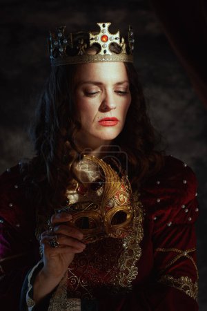 Foto de Reina medieval en vestido rojo con máscara veneciana y corona sobre fondo gris oscuro. - Imagen libre de derechos