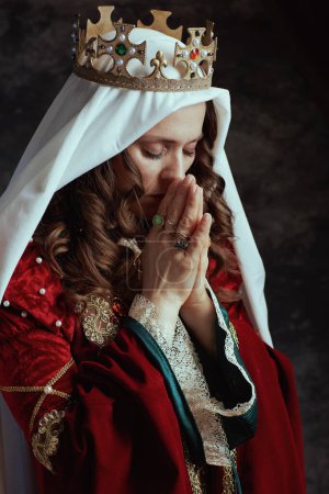 Téléchargez les photos : Reine médiévale en robe rouge avec voile et couronne priant sur fond gris foncé. - en image libre de droit