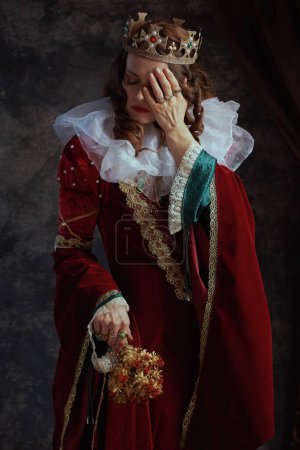 Photo for Unhappy medieval queen in red dress with dried flower, white collar and crown on dark gray background. - Royalty Free Image