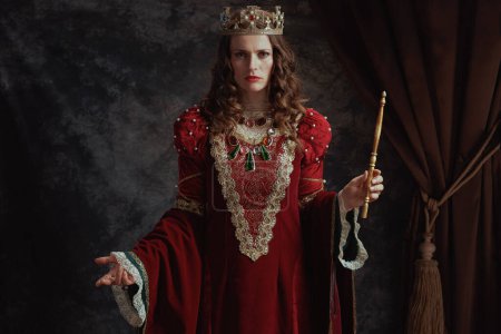 Foto de Reina medieval en vestido rojo con varita y corona sobre fondo gris oscuro. - Imagen libre de derechos