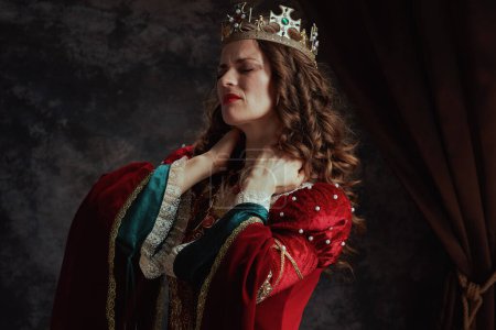 Foto de Cansada reina medieval en vestido rojo con corona con dolor de cuello sobre fondo gris oscuro. - Imagen libre de derechos