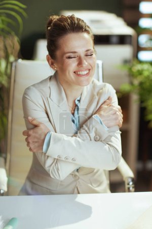 Foto de Feliz mujer moderna propietaria de una pequeña empresa en un traje de negocios ligero en la oficina verde moderna abrazándose. - Imagen libre de derechos
