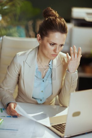 Foto de Mujer contable moderna infeliz en un traje de negocios ligero en la oficina verde moderna con documentos y computadora portátil. - Imagen libre de derechos