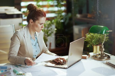 Foto de Moderna mujer contable de mediana edad en un traje de negocios ligero en la oficina verde moderna con documentos y computadora portátil. - Imagen libre de derechos