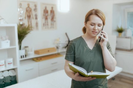 Photo for Healthcare time. concerned female medical massage therapist in massage cabinet with notebook talking on a smartphone. - Royalty Free Image