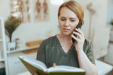 Photo for Healthcare time. concerned massage therapist woman in massage cabinet with notebook speaking on a smartphone. - Royalty Free Image