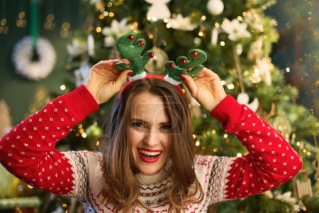 Photo for Christmas time. happy trendy woman with funny reindeer antlers headpiece hoop in traditional Christmas sweater near Christmas tree in the modern house. - Royalty Free Image