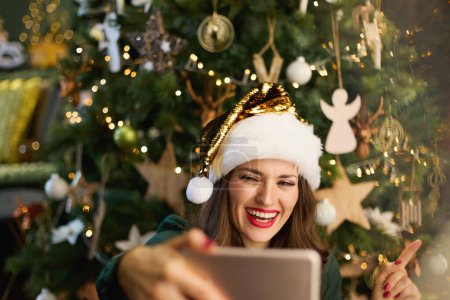 Foto de En Navidad. feliz mujer moderna con sombrero de Santa en vestido verde teniendo webcam reunión en un teléfono inteligente cerca del árbol de Navidad en el hogar moderno. - Imagen libre de derechos