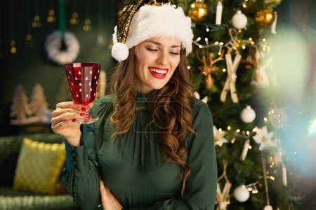 Foto de En Navidad. sonriente mujer de moda con sombrero de Santa y vidrio en vestido verde cerca del árbol de Navidad en la casa moderna. - Imagen libre de derechos