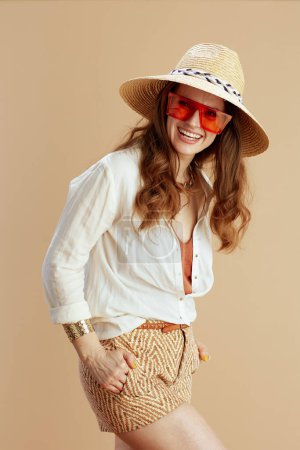Foto de Vacaciones en playa. feliz mujer moderna de 40 años en blusa blanca y pantalones cortos sobre fondo beige con sombrero de verano y gafas de sol. - Imagen libre de derechos