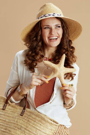Photo for Beach vacation. smiling stylish woman in white blouse and shorts on beige background with sea star, straw bag and summer hat. - Royalty Free Image
