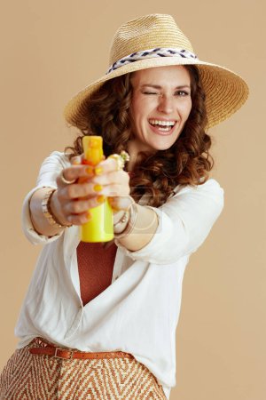 Photo for Beach vacation. smiling stylish middle aged housewife in white blouse and shorts isolated on beige background with spf and summer hat aiming as gun. - Royalty Free Image