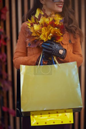 Photo for Hello october. happy trendy 40 years old woman in brown trench coat with shopping bags and autumn yellow leaves in the city. - Royalty Free Image