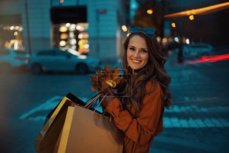Foto de Hola noviembre. mujer elegante feliz en gabardina naranja con bolsas de compras y hojas amarillas de otoño en la ciudad. - Imagen libre de derechos